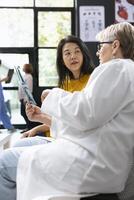 Inside a hospital lobby, medic does radiography scan interpretation with asian woman during checkup. Diverse people discussing about health evaluation and treatment guidance. Lifestyle awareness. photo