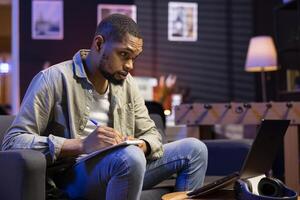 Young adult does his research by reading articles and taking notes on sofa in his living room, collecting information for a new project. Freelancer multitasking and studying data driven papers. photo