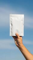Hand holding a blank white pouch against a clear blue sky, a clean slate for your brand design, Minimalist mockup of a sealed bag in front of azure, perfect for packaging visuals photo