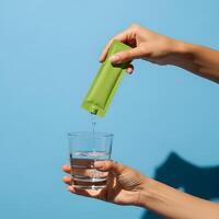 Green packet being poured into glass of water against blue background, Creating a refreshing hydration moment with a convenient green packet drink photo