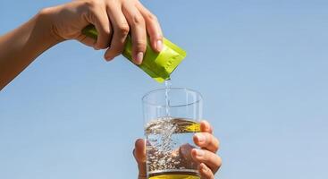 Person pouring electrolyte powder from a green packet into a glass of water, Hydration with Electrolytes in Glass Against a Blue Sky photo