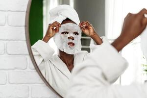 Woman applies a facial mask while looking in the mirror at home during a self-care routine photo
