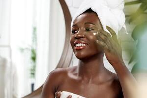 Woman applying moisturizer in a bright bathroom with a towel on her head during skincare routine photo