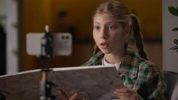 Clever Caucasian schoolgirl little girl teenager kid child influencer blogger talking online streaming reading book record blog podcast audio read literature mobile phone tripod camera at home photo