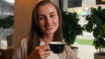 Relaxed Caucasian woman drinking coffee in cafe girl female sitting at table in cafeteria visitor drink tea croissant morning breakfast pause in restaurant looking at camera smiling happy satisfied photo