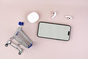Shopping cart with a smartphone and white wireless headphones on a pink background. photo
