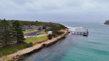 Aerial view of Port Campbell's pier and coastline, showcasing lush greenery, ocean waves, and overcast skies captured by a drone video