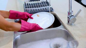 Hands in pink gloves wash dishes under running water in a bright kitchen. The sequence captures the cleaning process with close-up shots video