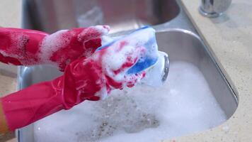 Hands in pink gloves wash a glass with a sponge in a kitchen sink, surrounded by soapy water video