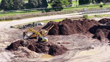 Drone captures an excavator moving soil and mulch in a sunny, industrial landscape with greenery in the background video