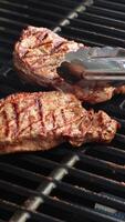 Beef steaks cooking on a grill with tongs turning them. Sunlit outdoor setting enhances the appetizing appearance video