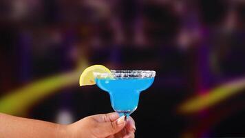 A hand holds a blue margarita cocktail with a lemon slice, set against a colorful, blurred background video
