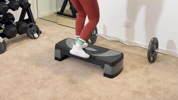 A woman performs step exercises with dumbbells in a home gym setting, focusing on fitness and strength video