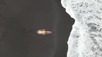 Top down view of woman in yellow swimsuit lying on volcanic black sand beach with ocean waves in Bali, Indonesia video