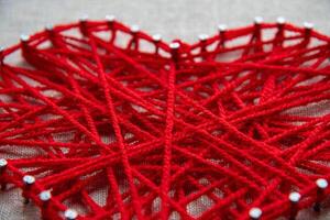 Colorful and intricate string art displays a heart shape formed by intertwined red yarn wrapped around small nails on a textured background. photo