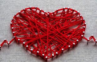 Craft string art in shape of heart. Red woolen heart, symbol of love, made of red wool yarn threads tangled over metal nails on canvas background photo