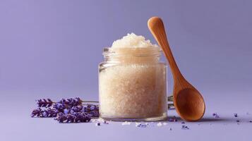 Ceramic jar filled with sugar scrub and wooden spoon beside lavender flowers on a purple background photo