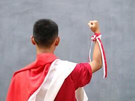 Young boy in red shirt with Indonesian flag on his shoulder, his fist raised upwards, during Indonesia's Independence day celebration, back view photo