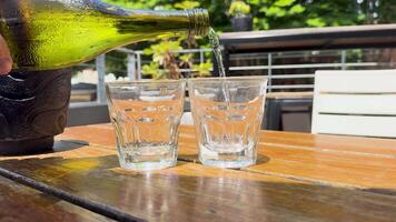 A bottle pours water into glasses on a sunlit outdoor table, creating a refreshing scene with vibrant greenery video