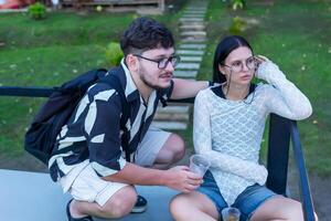 Man and woman at sunset. Young couple is seated on terrace, enjoying drinks while surrounded by lush green grass and wooden path in background, creating relaxed and casual atmosphere for socializing photo