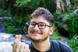 Young man with glasses is smiling and making heart gesture with his hands, surrounded by vibrant greenery and natural elements, creating joyful and friendly atmosphere. Traveler with backpack photo