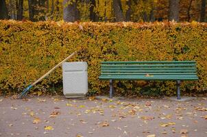 In the autumn park, there is a bench, next to an urn and a rake for cleaning leaves. High quality photo