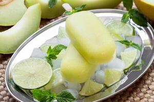 Melon lime sorbet ice cream on silver plate with ice cubes, lime pieces, mint leaves near melon slices, lime on a straw napkin background. Melon and lime sorbet ice cream popsicles. Horizontal. photo