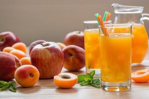 Apricot peach apple juice in glasses with ice near jar of juice and scattered apricots, peaches, apple mint on light wooden background. Apricot peach apple juice with ice. Horizontal. Daylight. photo