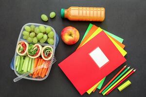 Box with wraps with vegetables, grapes, apple, juice bottle, colorful notebooks and pencils arranged neatly on black background for school lunch preparation. photo