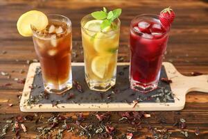 Three tall glasses of iced drinks placed on a wooden tray feature bright colors and fresh garnishes. Ideal for warm days, these refreshments showcase a variety of flavors and ingredients. photo