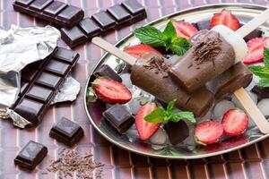 Chocolate vanilla ice cream on a silver platter with ice and chocolate pieces, mint, strawberry near pieces of chocolate on a chocolate background. Chocolate vanilla ice cream popsicles and stawberry. photo