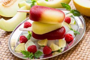 Column of melon lime raspberries sorbet ice cream on silver plate with ice cubes, lime, mint, raspberries near melon slices on straw napkin background. Melon lime raspberries sorbet ice cream. Close. photo