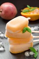 Column of mango sorbet sundae mix ice cream behind mango and cutted mango near ice cubes on a black background. Mango sorbet sundae mix ice cream popsicles. Vertical. photo