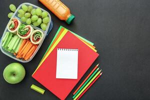 School supplies and lunch box with tasty rolls, cucumbers, carrots, grapes, apple, bottle of juice on black background, back to school concept with copy space. Flat lay. Top view. photo