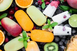 A vibrant array of berry flavored ice cream bars laid out flat, surrounded by slices of fresh fruits including melons, oranges, kiwis, and mint leaves for decoration. photo