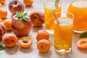 Apricot peach apple juice in glasses with ice near the jar of juice and scattered apricots, peaches, apple, mint on wooden background. Apricot peach apple juice with ice. Horizontal. photo