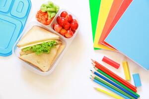 Lunchbox filled with sandwiches, cucumber, carrots and cherries placed next to vibrant stationery, including notebooks and colored pencils, ideal for school activities on white backgrond. photo