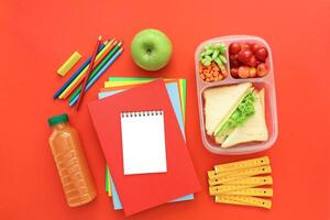 Colorful lunchbox filled with fruits, vegetables and sandwich sits beside green apple, school supplies, and drink on bright red surface. Ideal setting for preparing healthy meals. photo