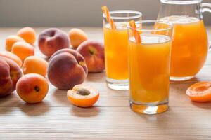 Glasses filled with bright apricot peach juice are placed on a wooden table, surrounded by fresh fruits and a pitcher. The warm sunlight illuminates the scene. photo