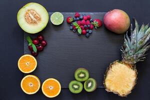 Rectangle from laid out fruit pineapple, mango, kiwi, oranges, lime, melon and berries cherries, strawberries, blueberries, raspberries in middle copy space on black background. Top view, flat lay photo