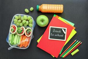 School supplies and lunch box with tasty rolls, cucumbers, carrots, grapes, apple, bottle of juice on black background, back to school concept. Flat lay photo