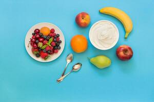 Greek yogurt around orange, banana, pear, peach, apple, plate with strawberries, raspberries, blueberries and two spoons on light blue background. Horizontal. Top view. photo
