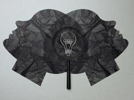Two symmetrical black silhouettes of faces, in the center between them is a black magnifying glass, a light bulb painted in white photo