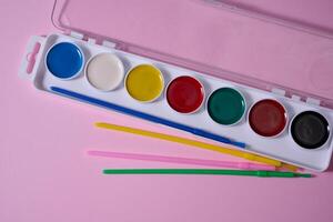 Set of watercolor paints in a plastic case with multi-colored brushes is located on a pink background. Top view photo