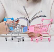 Person's hands holding unfolded stacks of hundred dollar bills in front of two miniature shopping carts standing on white surface. photo