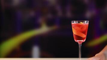 A Negroni cocktail is placed on a bar counter with vibrant lighting and a blurred background video