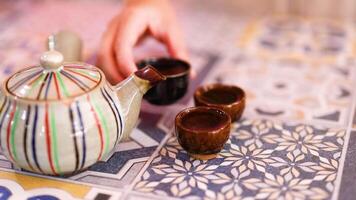 A hand pours tea from a striped teapot into small cups on a patterned tablecloth, creating a calming atmosphere video