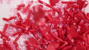 Red heart-shaped candies and sprinkles cascade onto a white surface, creating a vibrant, sweet composition video