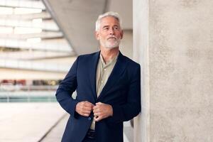 Senior businessman adjusting suit while leaning on concrete wall photo