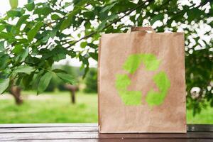 Blank paper bag stands on a wooden table against the background of green leaves. photo